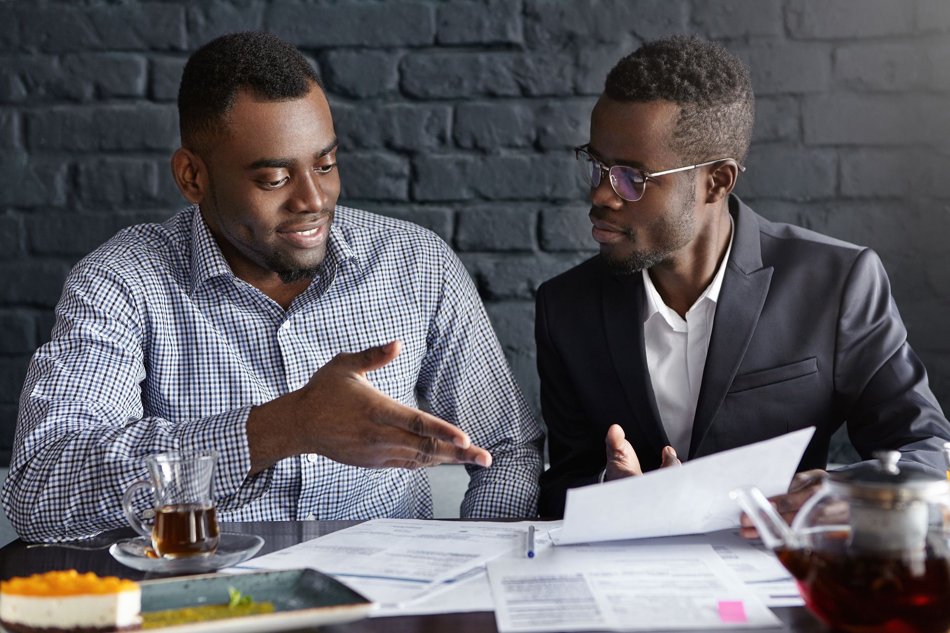 Two men reviewing documents at a table. One gestures, and the other looks on while holding papers.