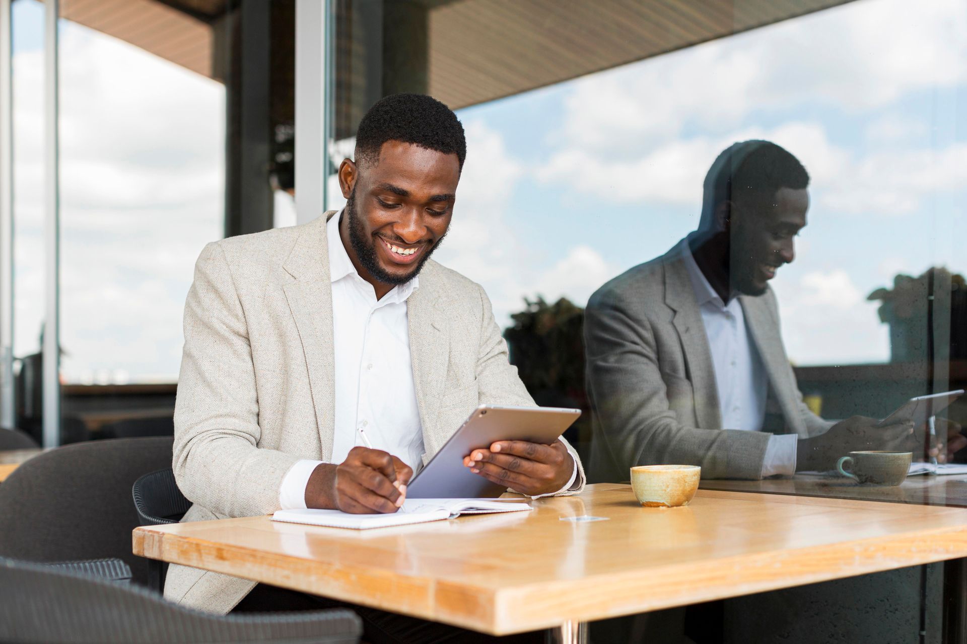 Man in a blazer smiles while using a tablet at a table with a cup, reflected in glass.