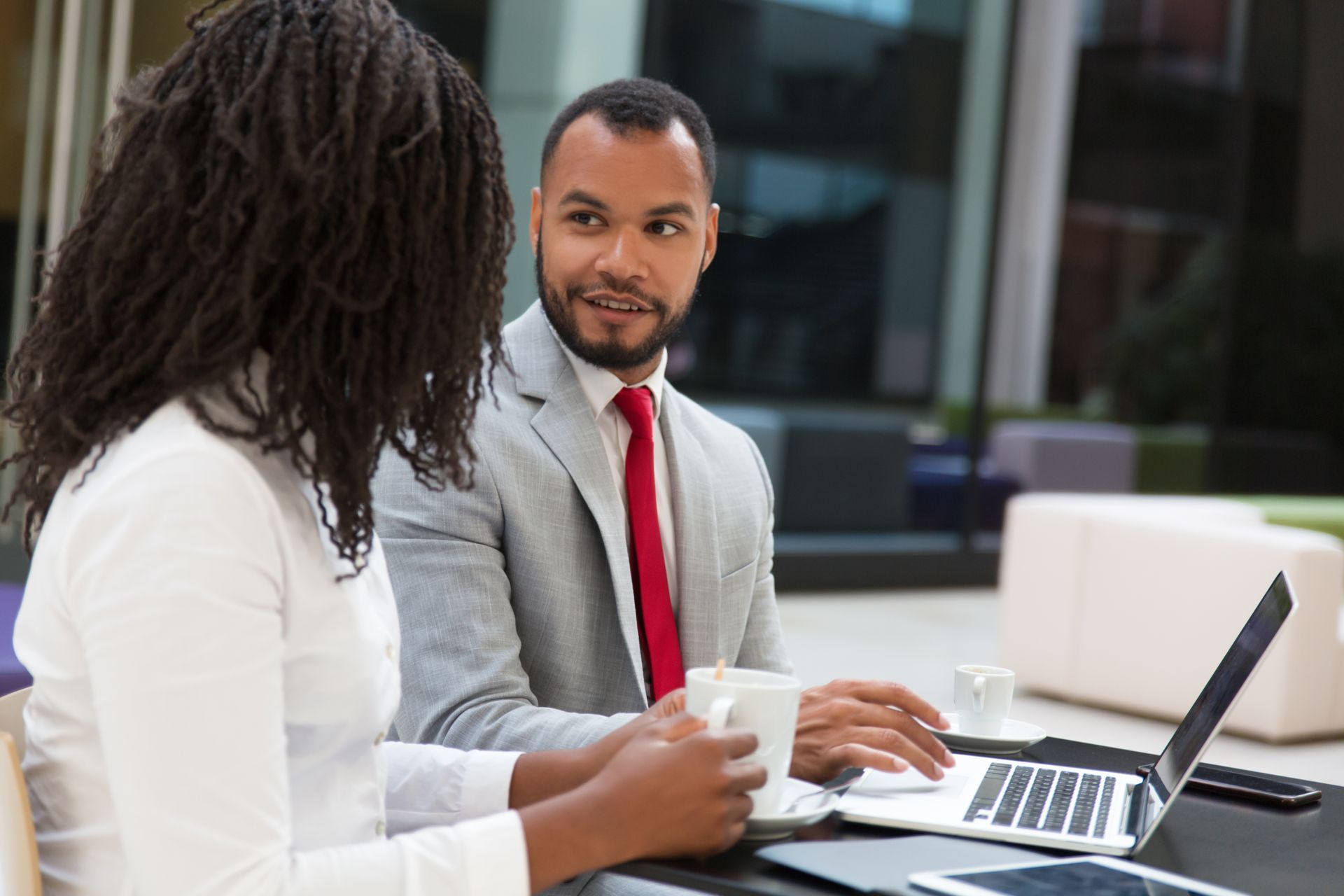 Man in suit, red tie, speaking to person with long hair, looking at laptop.