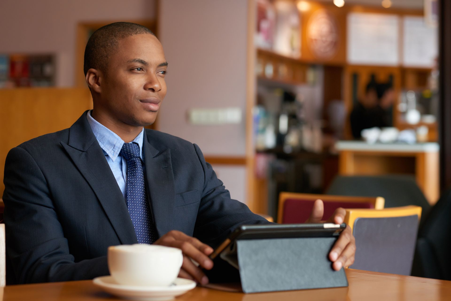 Man in a suit at a table, holding a tablet and coffee cup, looking thoughtfully to the side in a cafe.
