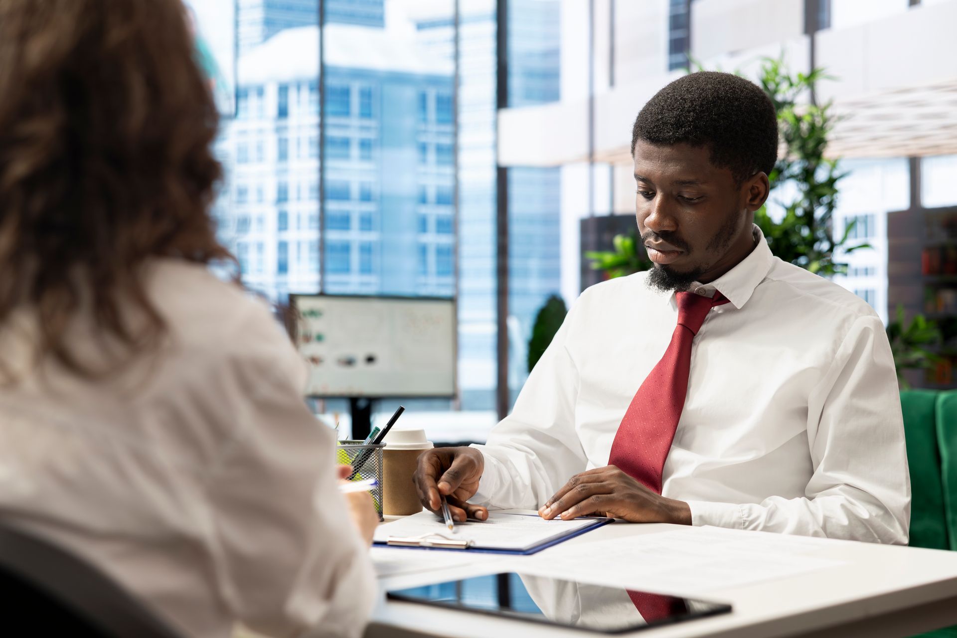 Man in white shirt and red tie writing at a desk, facing another person. Office setting.