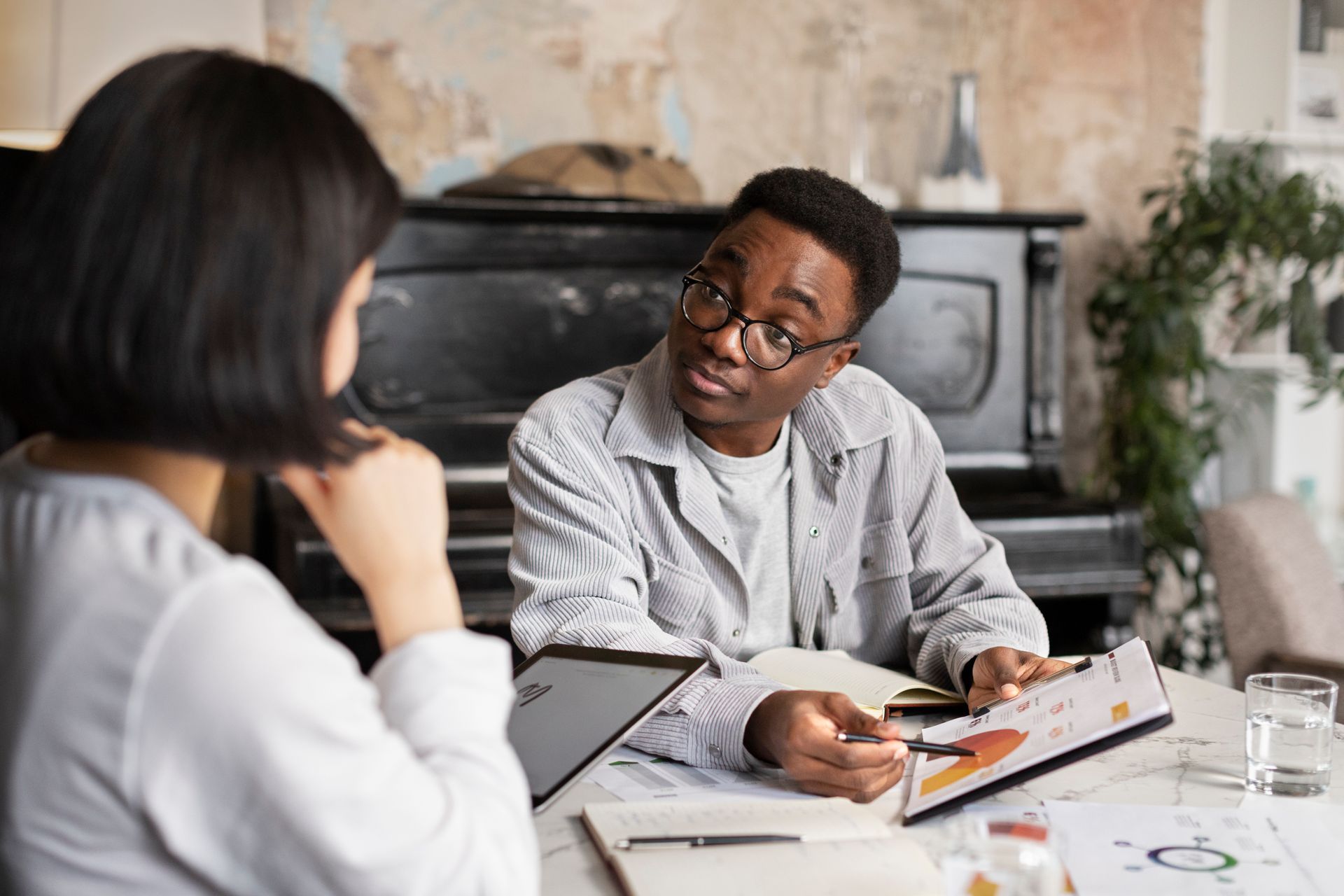 Man showing a document to a woman at a table, discussing work. A piano sits in the background.