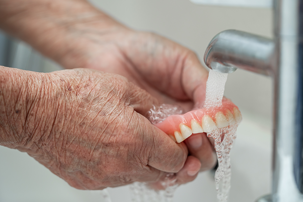 hands of an older person washing off their dentures in the sink under the water