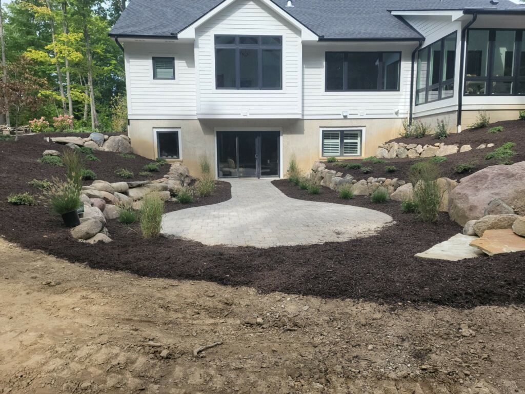 A gardener in an orange shirt and green overalls kneeling to trim a vibrant green hedge on a sunny day.