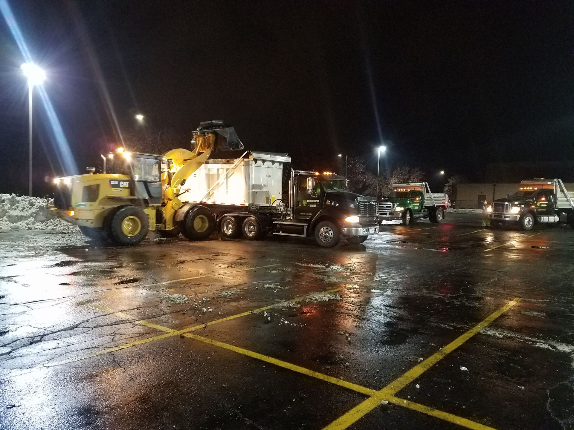 A white skid-steer loader with a yellow snowplow blade clearing snow in a parking lot near a UPS trailer.