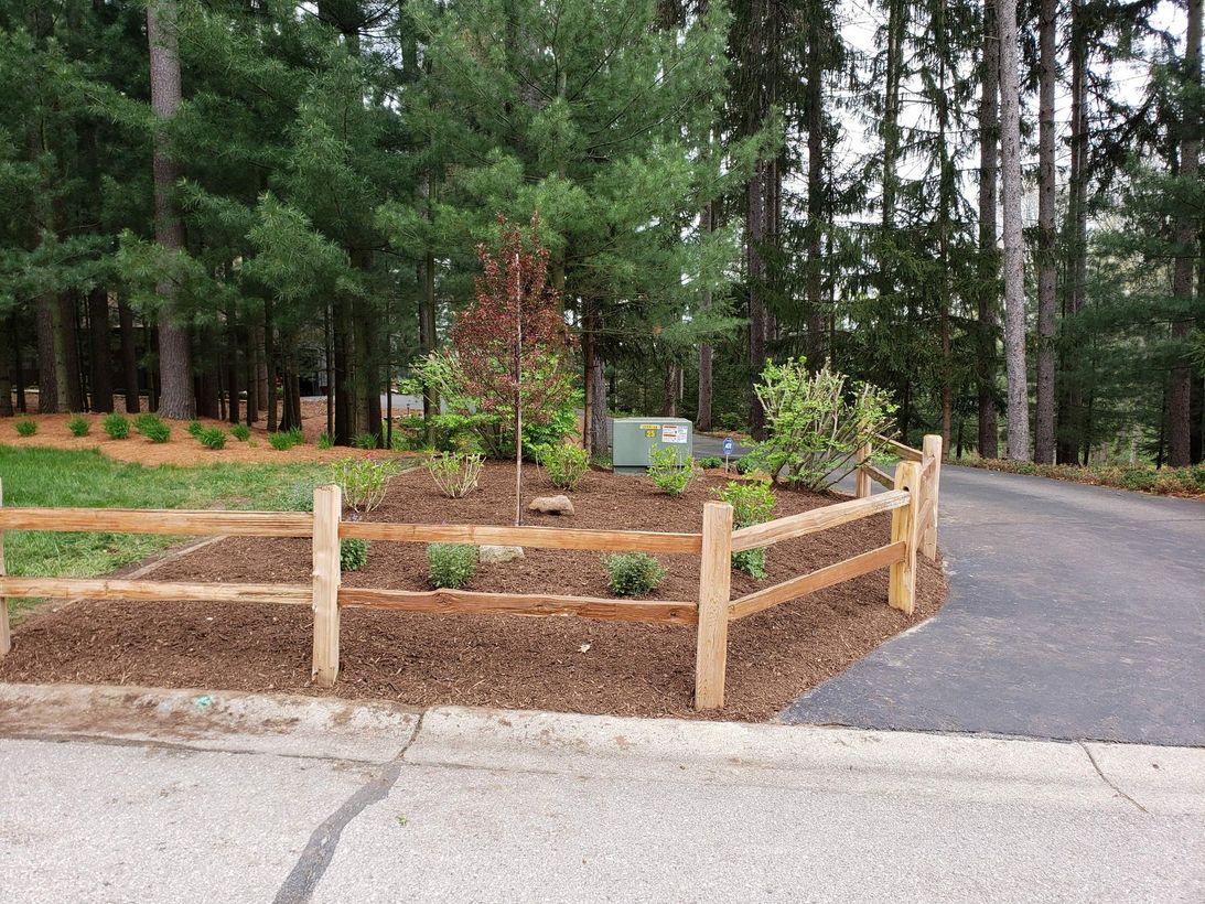 A gardener in an orange shirt and green overalls kneeling to trim a vibrant green hedge on a sunny day.