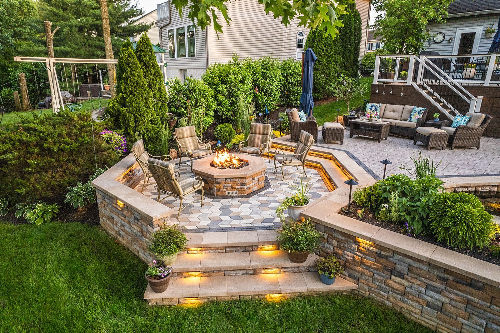 A sunken stone fire pit patio with built-in stairs, ambient lighting, and outdoor seating in a landscaped backyard.