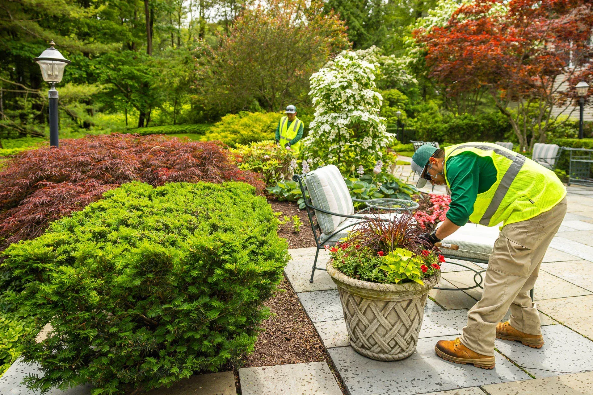A landscape worker in a high-visibility vest tends to a potted plant on a patio, with another worker visible in the garden.