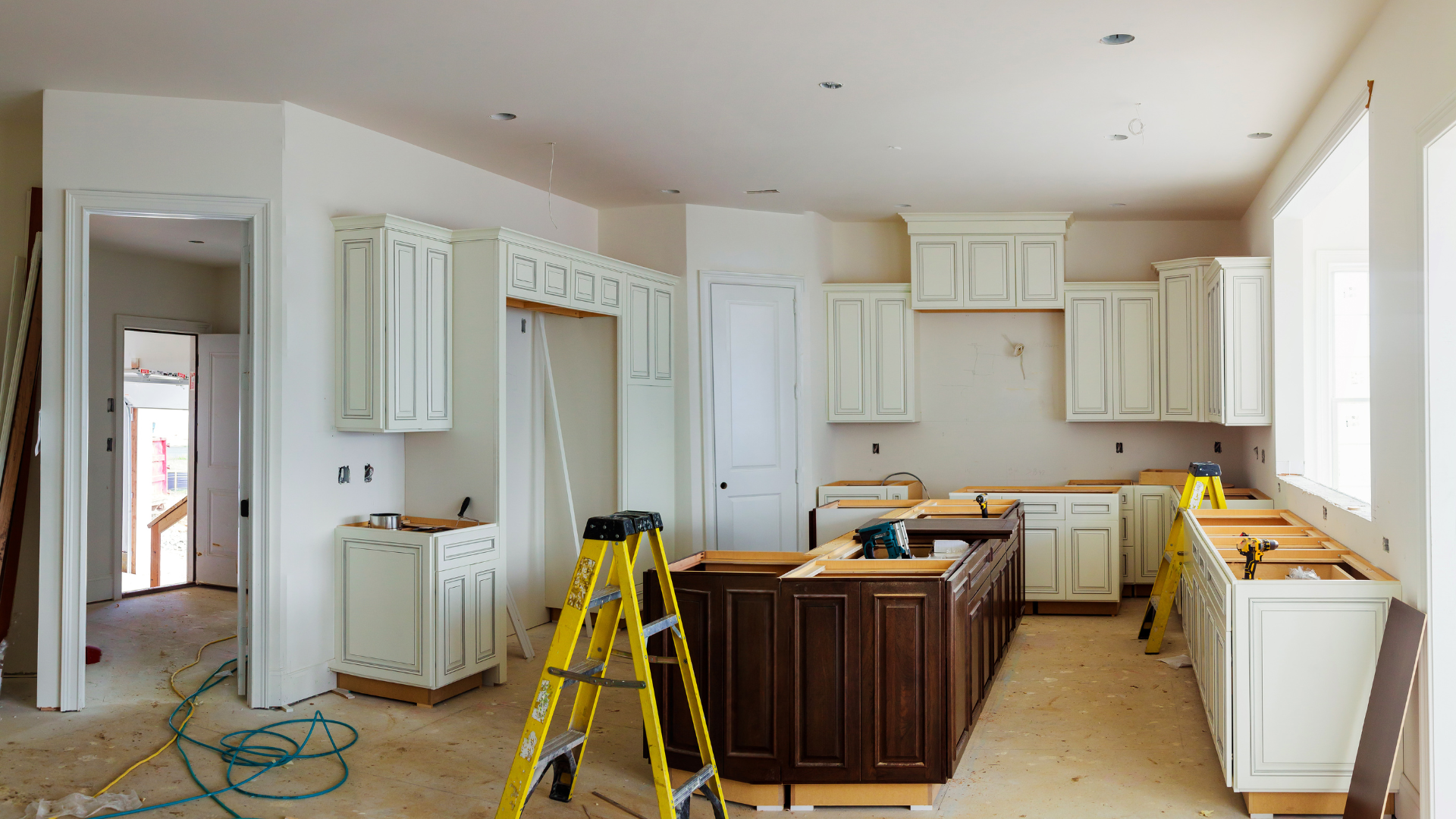 Bright kitchen under renovation with white cabinets, island, and a yellow ladder on a covered floor.