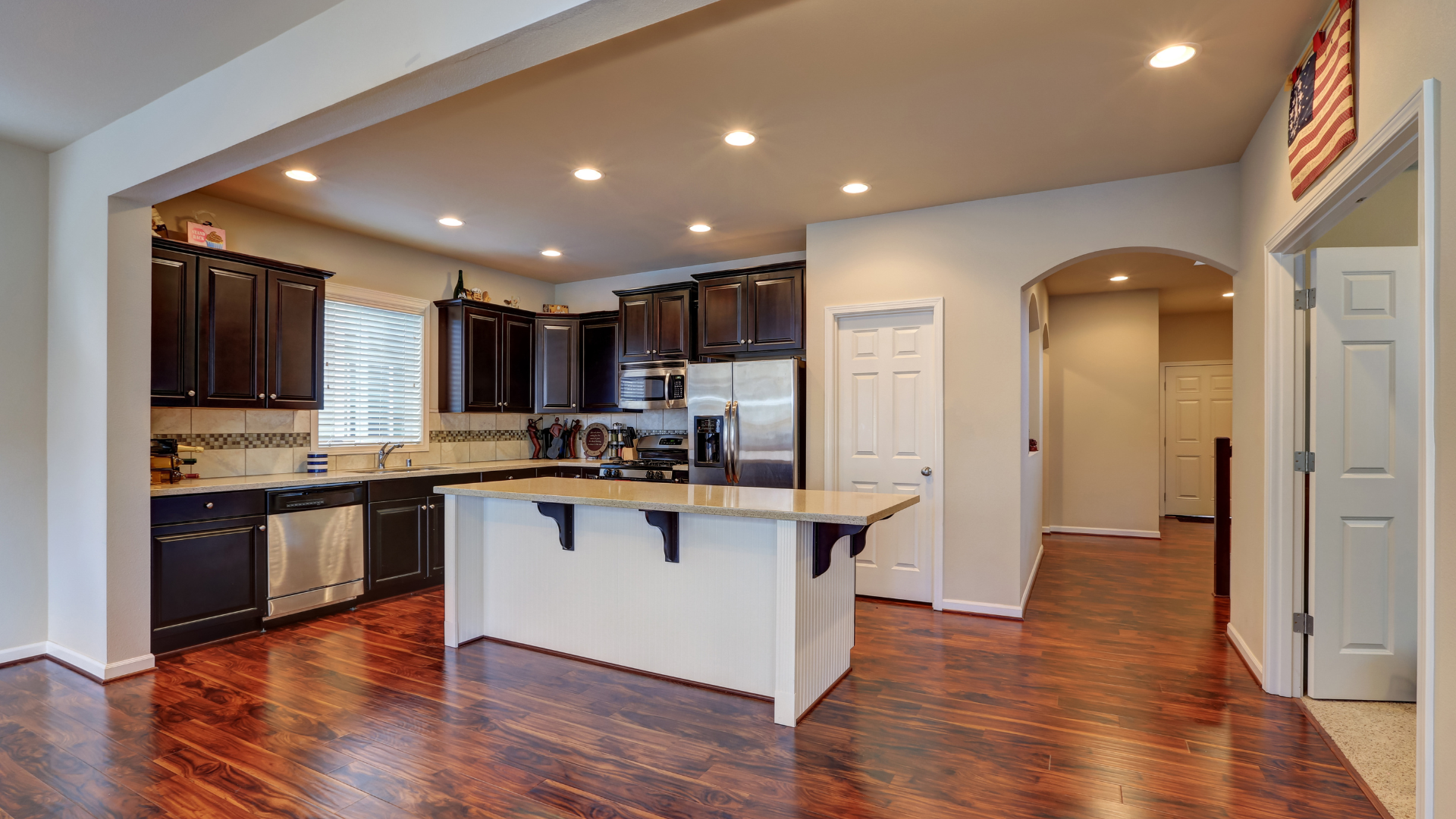 Modern kitchen with dark wood cabinets, white island, and hardwood floors, viewed into an adjoining hallway