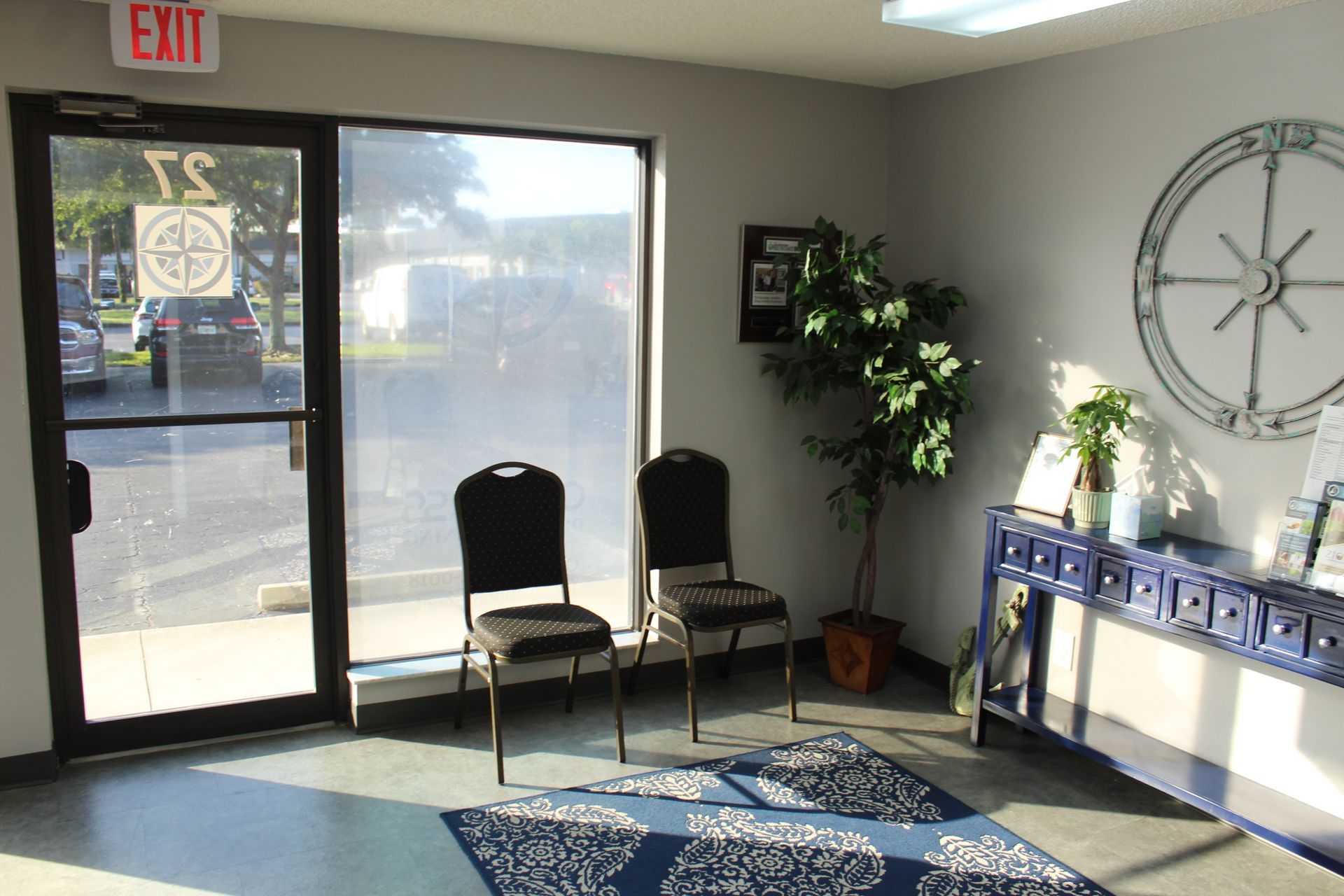 A waiting room with two chairs , a plant , and a compass on the wall.