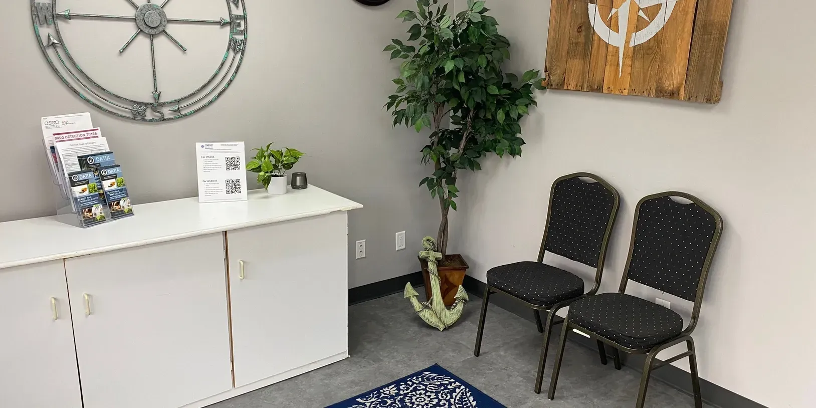 A waiting room with two chairs , a plant , and a compass on the wall.