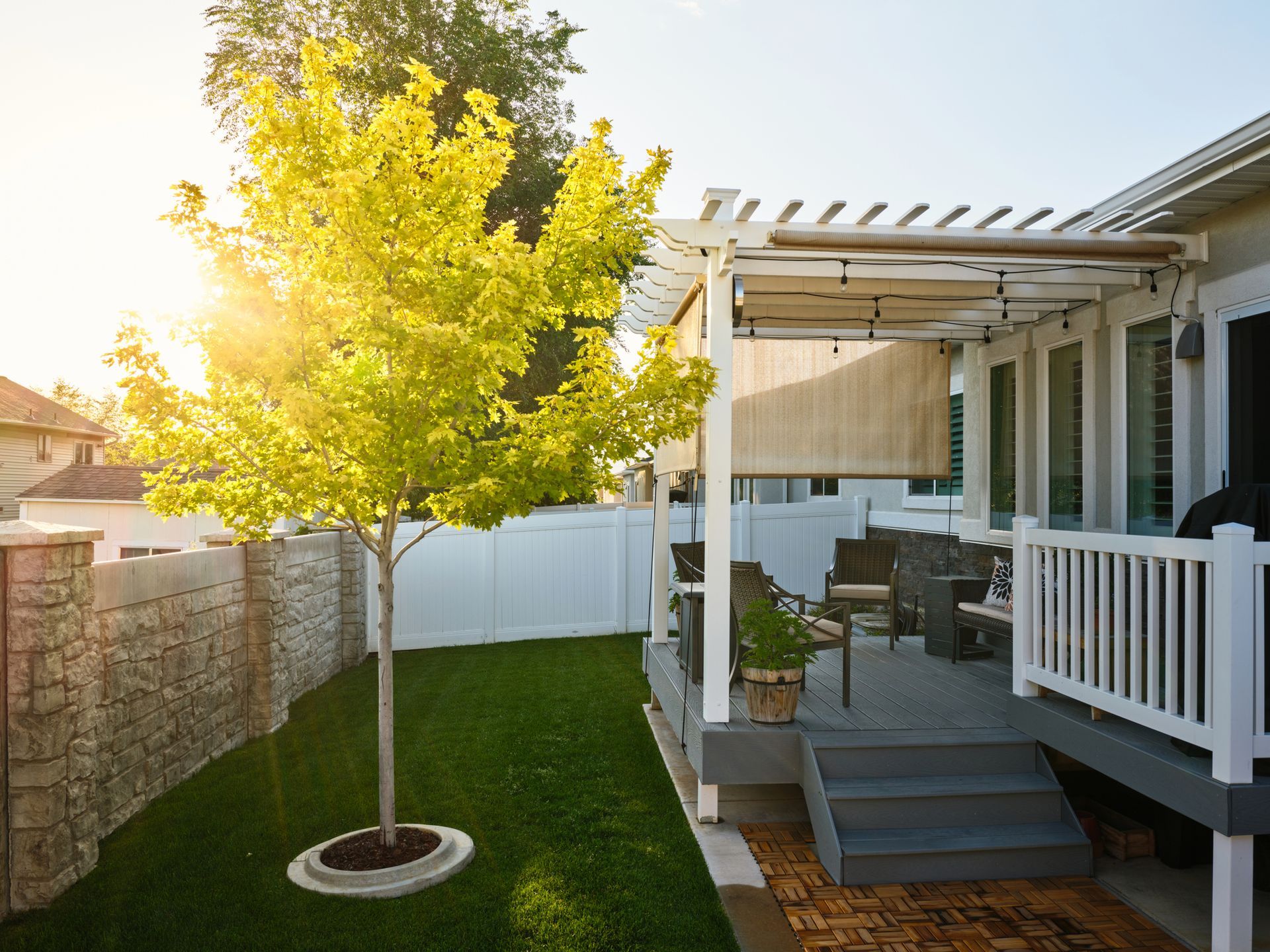 Motorized shades partially lowered over patio pergola with outdoor furniture and sunny yard.