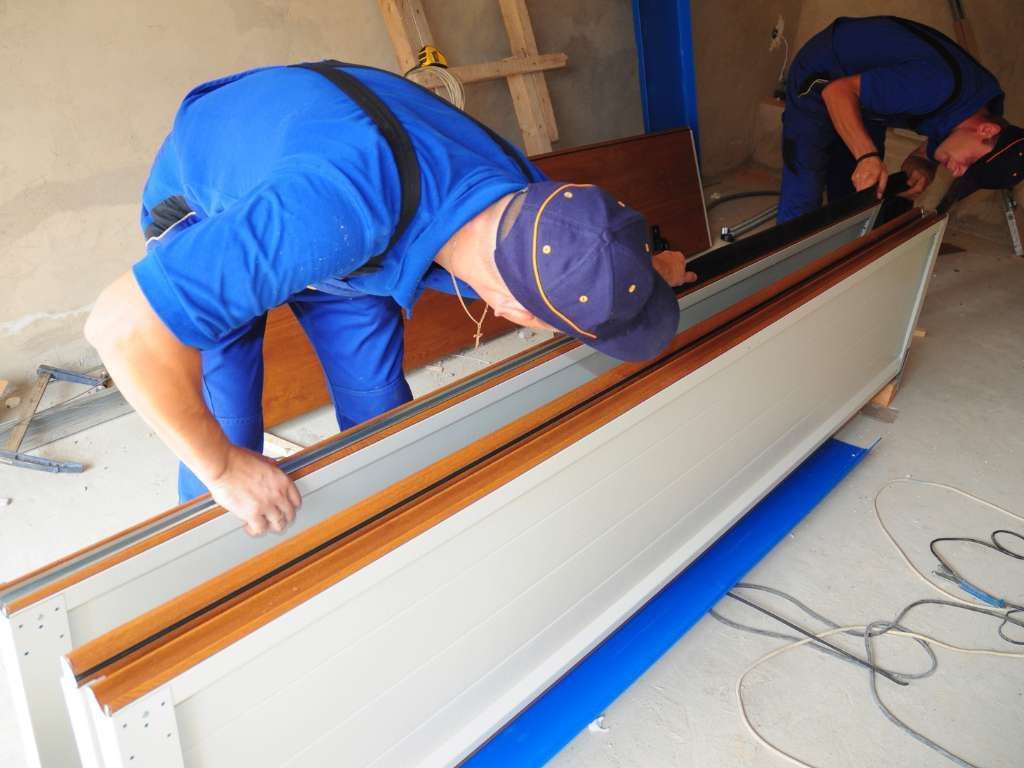 Two workers in blue coveralls installing a door, indoors. One bends over the frame, the other works in the background.