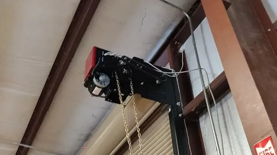 Black electric chain hoist mounted on a vertical support inside a warehouse.