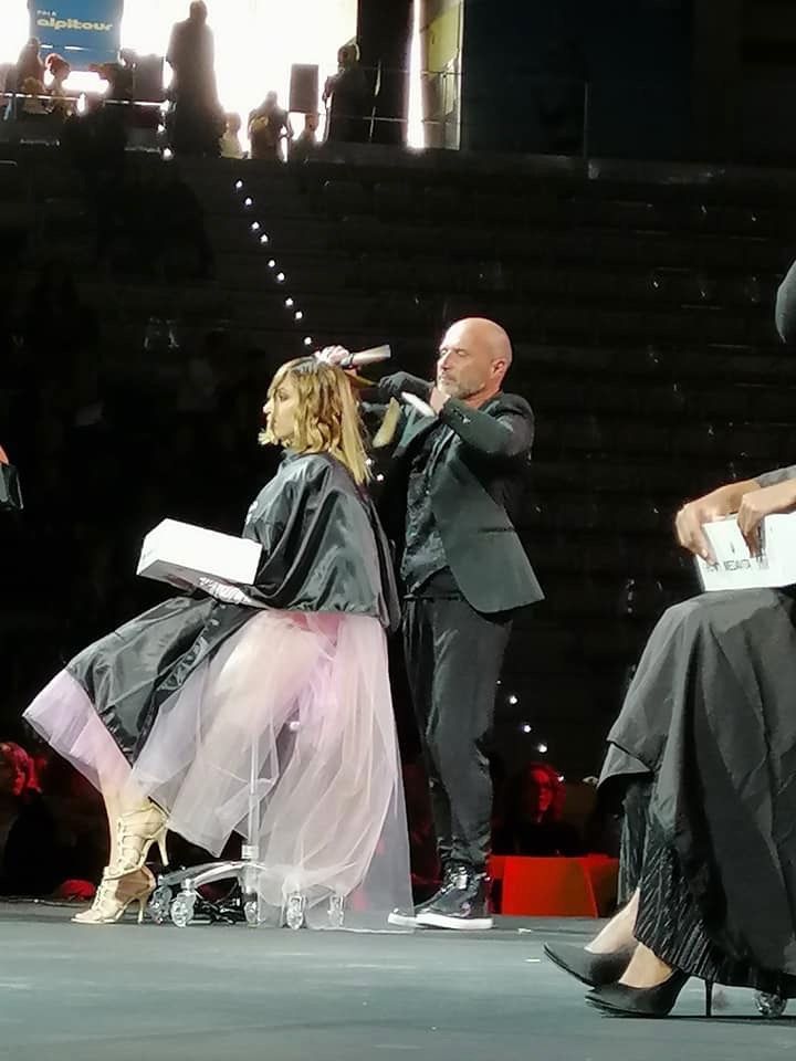 Una donna con una gonna rosa si sta facendo tagliare i capelli da un uomo in giacca e cravatta su un palco.