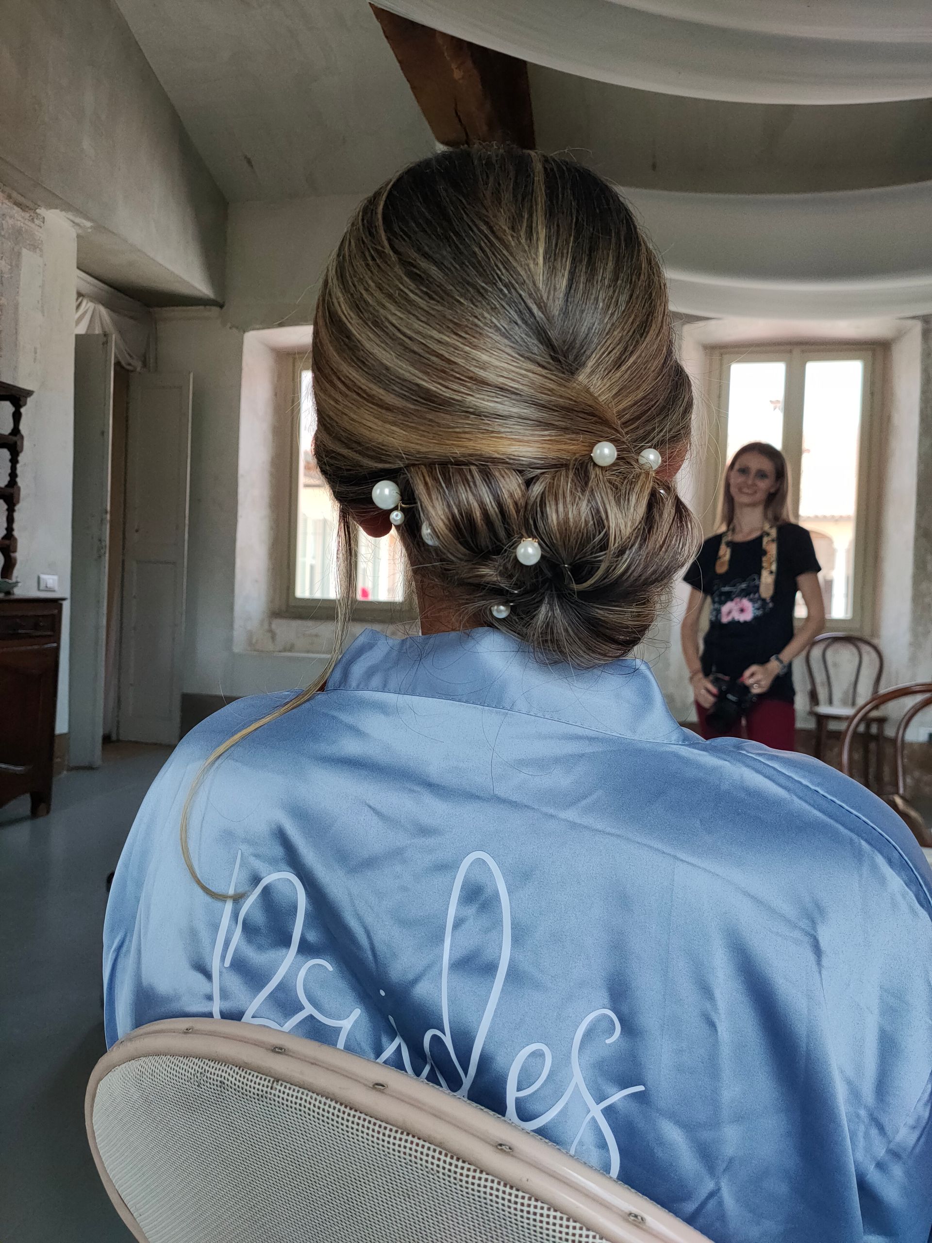 Una donna con una tunica blu è seduta su una sedia con i capelli raccolti in uno chignon.