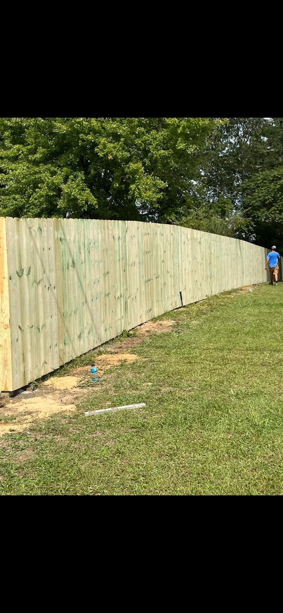 A man is standing next to a long wooden fence in a grassy field.