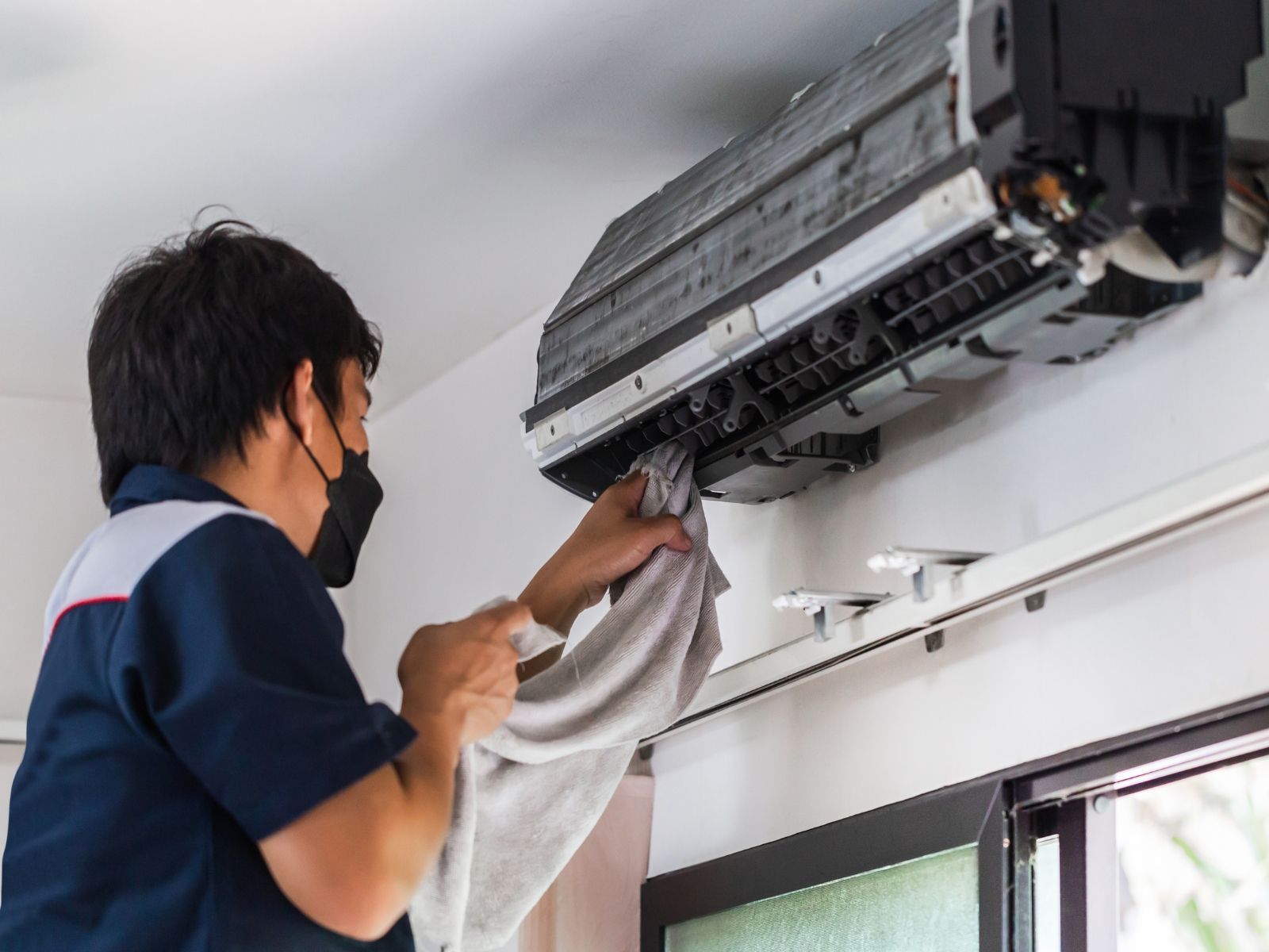 A man wearing a mask is cleaning an air conditioner with a towel.