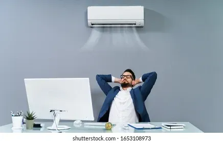 A man is sitting at a desk in front of a computer under an air conditioner.