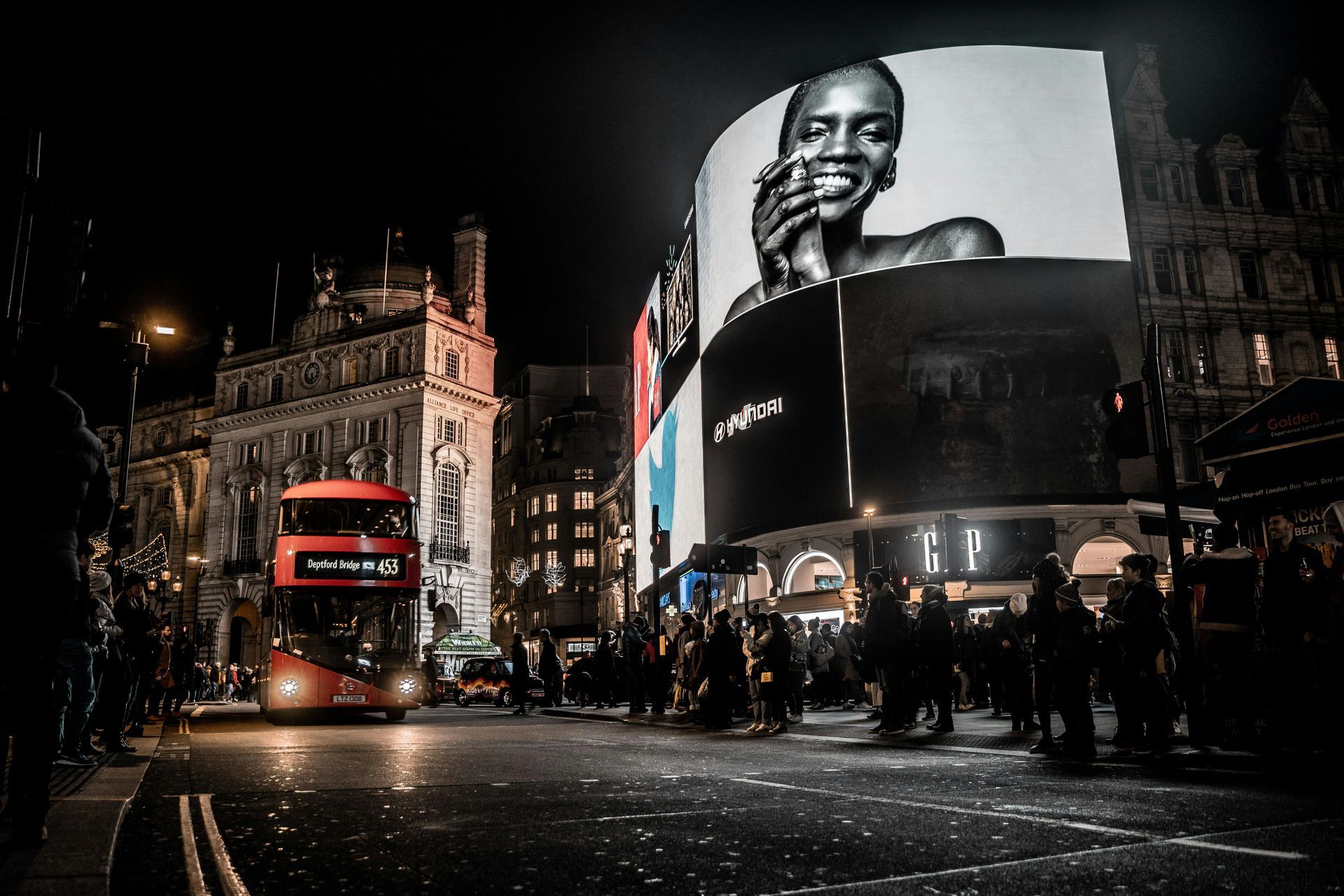 A double decker bus is driving down a city street at night in front of a large billboard.