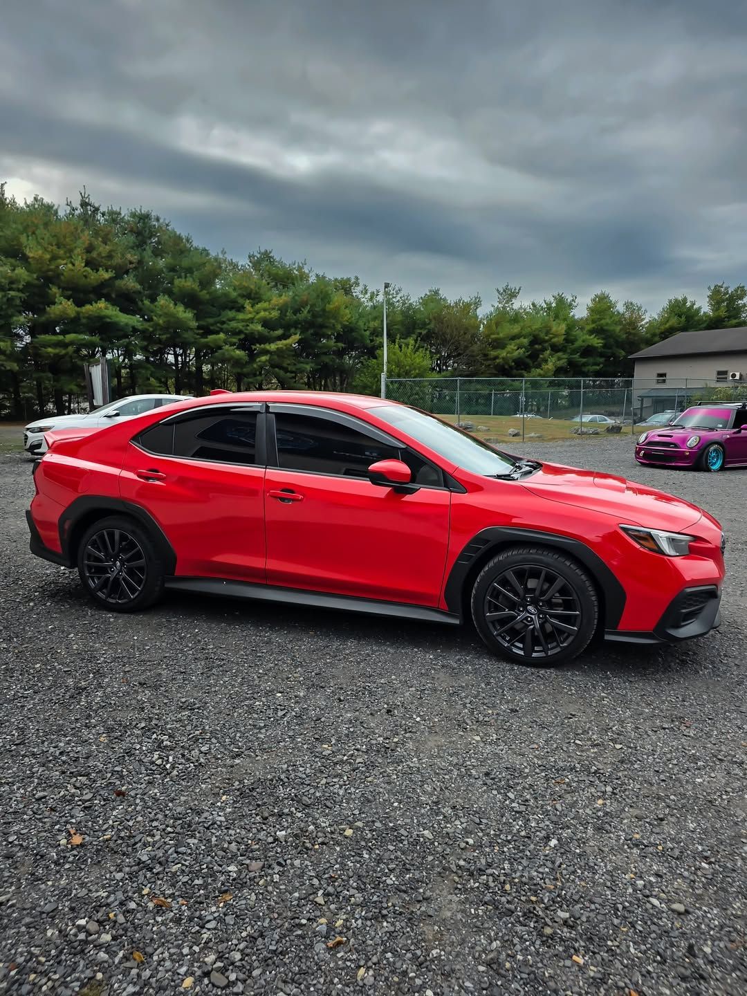 Red Subaru car with black wheels parked on gravel. Overcast sky.