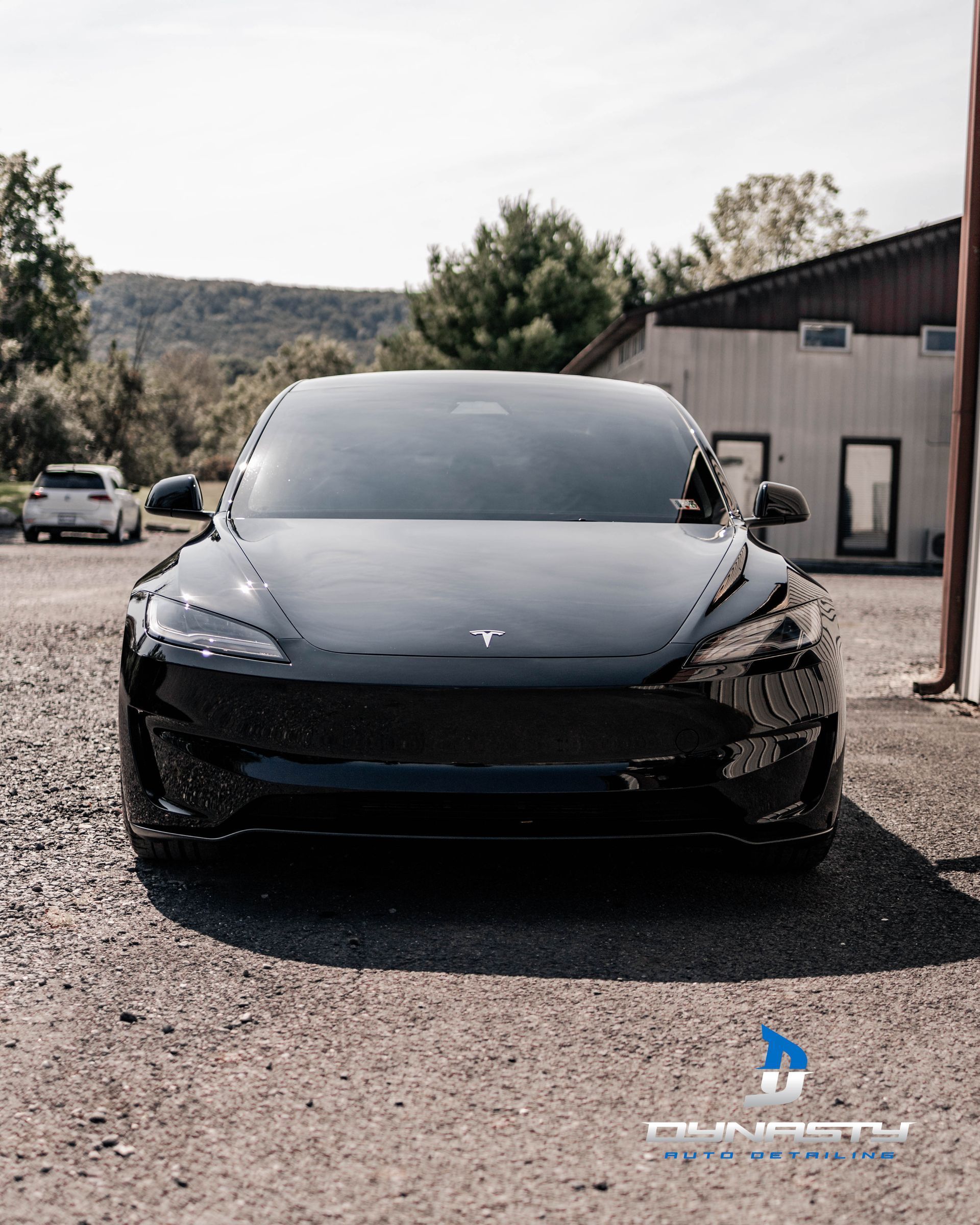 A black tesla model 3 is parked in a gravel lot in front of a building.