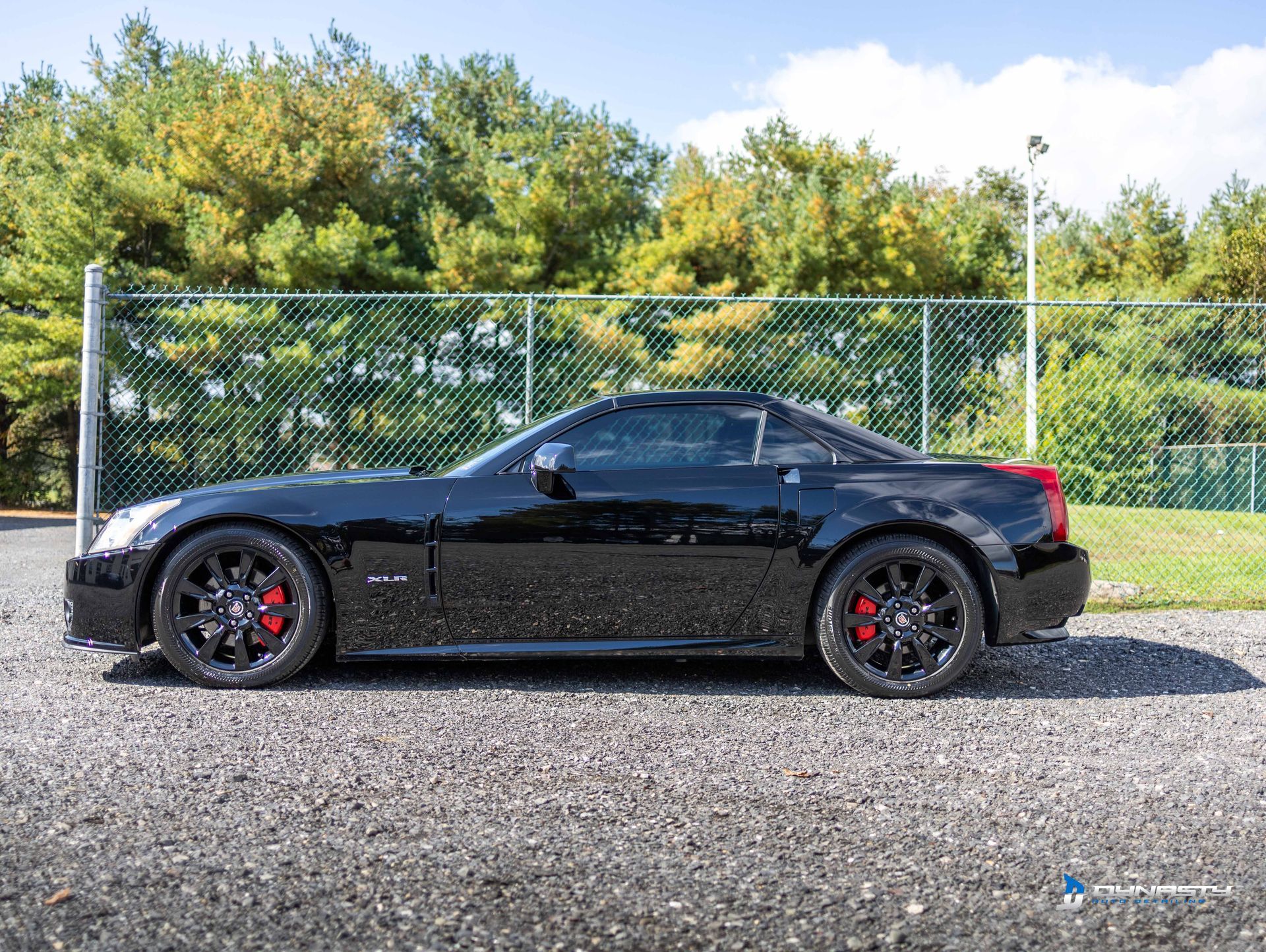 A black sports car is parked in a gravel lot next to a chain link fence.