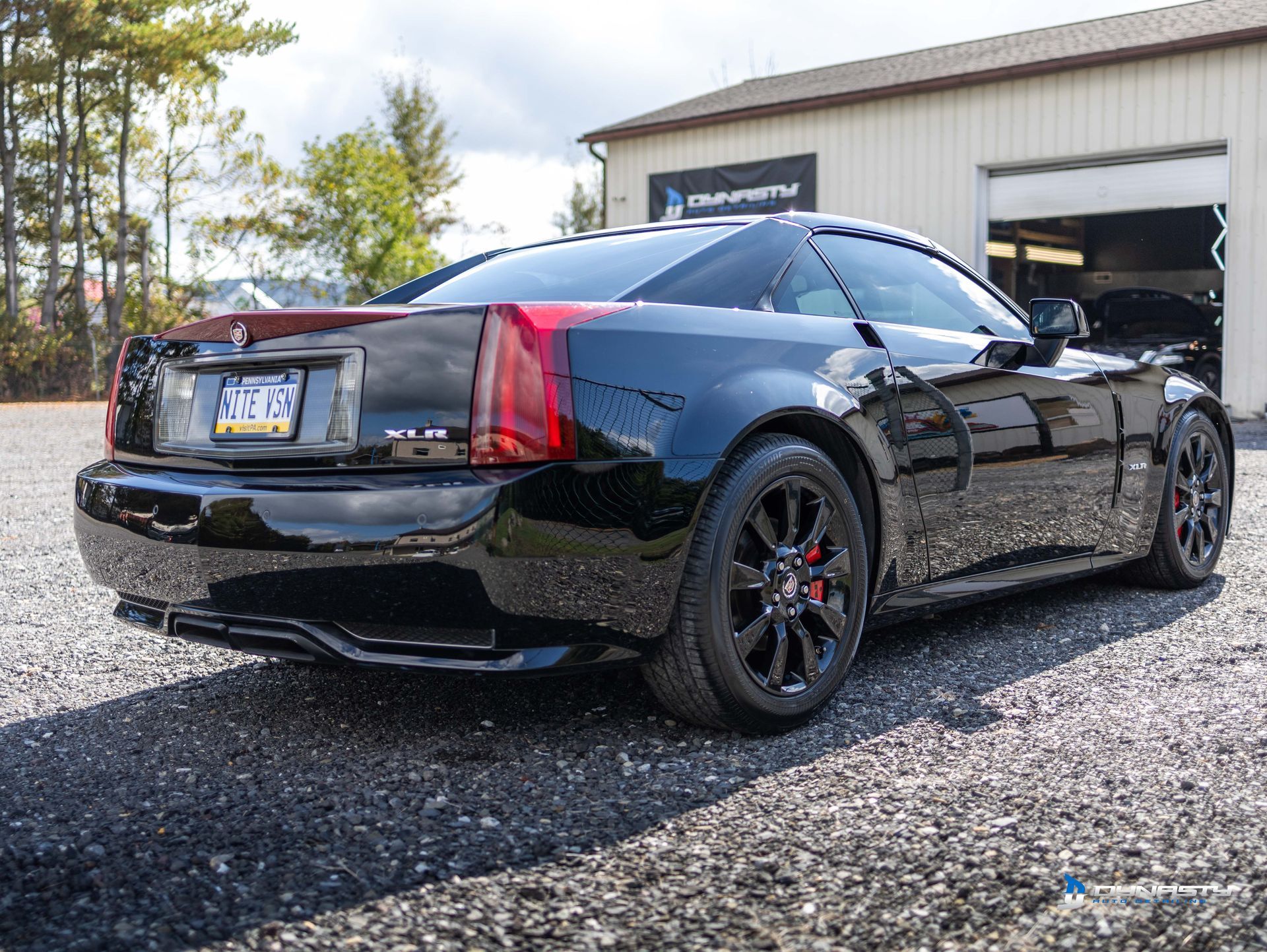 A black car is parked in a gravel lot in front of a garage.