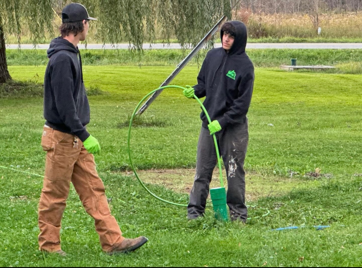 Two men are standing in the grass holding a hose.
