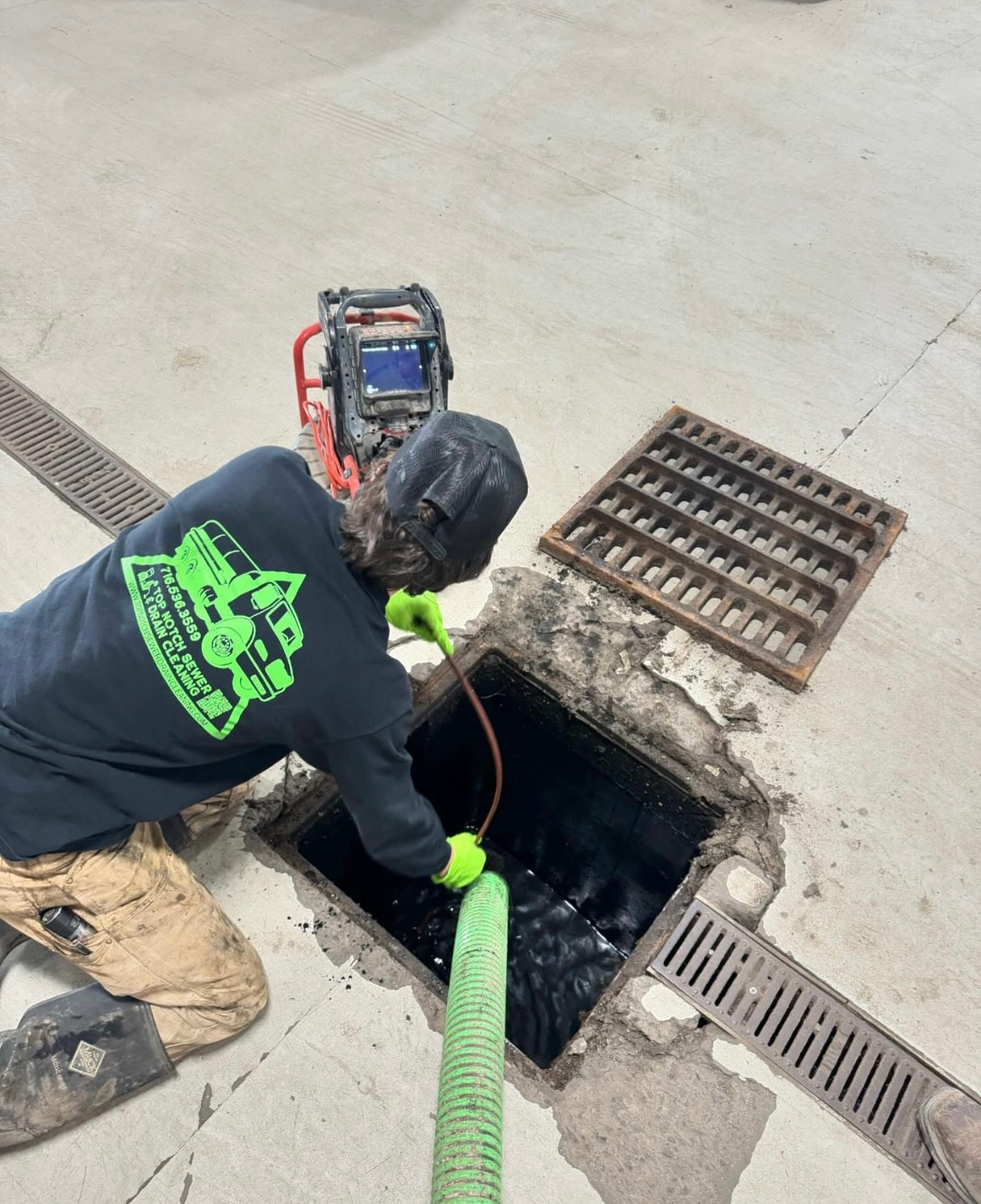A man in a black shirt with a green logo on the back is working on a drain