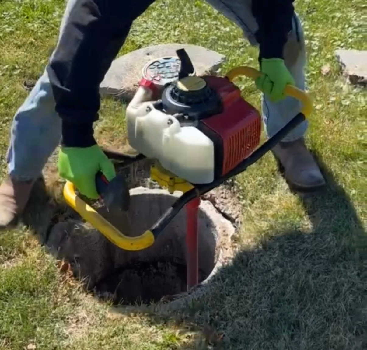 A man wearing green gloves is using a drill to dig a hole in the ground