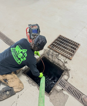 A man in a black shirt with a green logo on the back is working on a drain