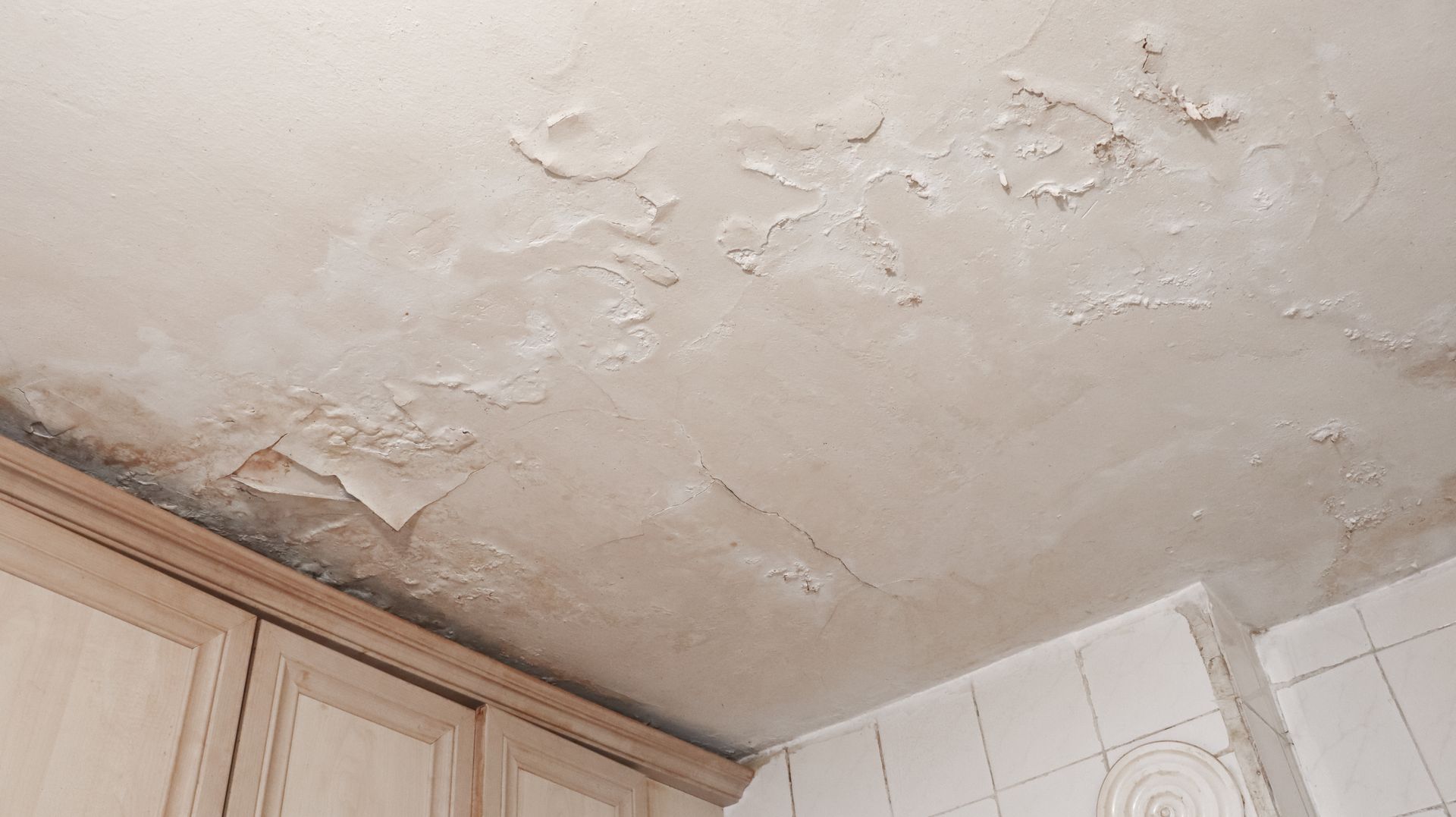 A kitchen ceiling with severe water damage, showing peeling paint, bubbles, and dark, damp patches above wooden cabinets.
