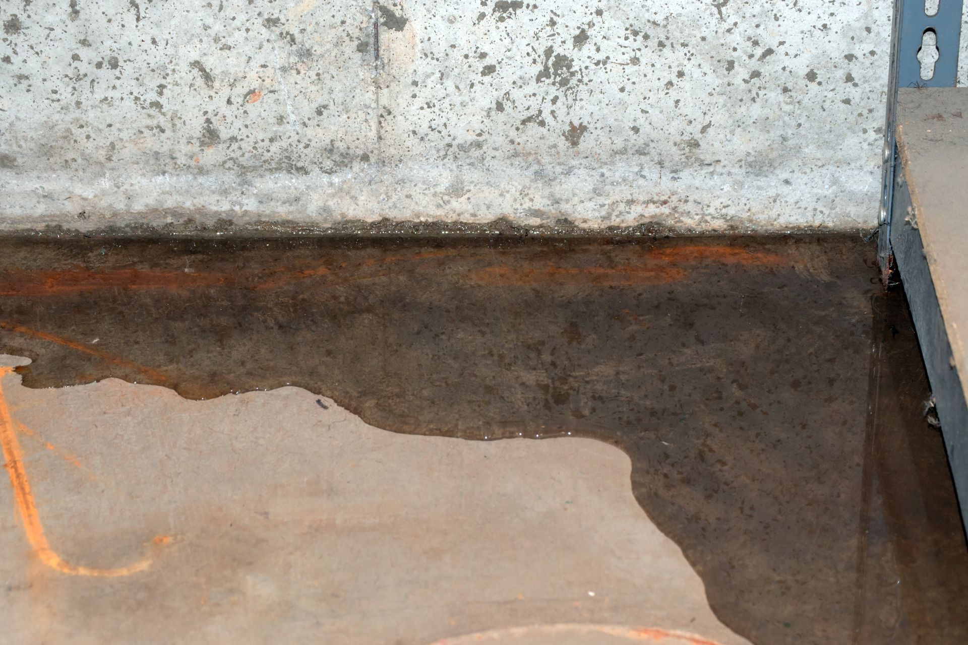 Water pooling on a concrete basement floor against a foundation wall with signs of rust or water damage.