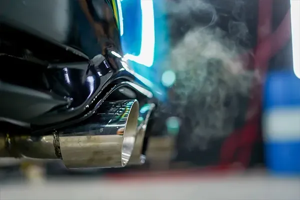Steam rising from a motorcycle exhaust pipe in a close-up shot, with blurred blue and red lights in the background