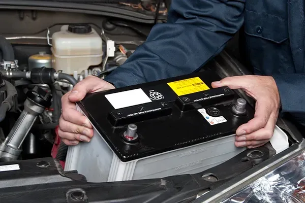 Mechanic installing a black car battery in an engine bay, with labels visible.