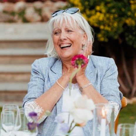 Woman with grey hair wearing sunglasses, black blazer, white pants, and pearl necklace. Standing in a parking lot.