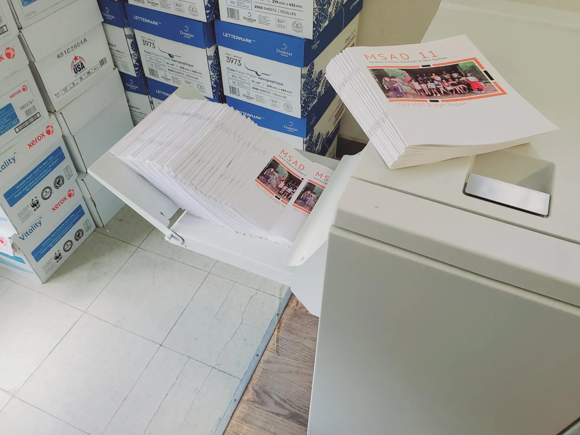 Stacks of printed documents with color photos and stamps on a white shelf, next to a printer and boxes.
