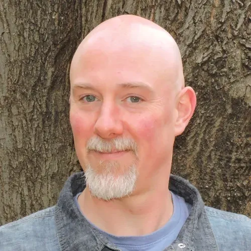 Bald man with goatee smiles in front of a tree, wearing a gray shirt over a blue shirt.