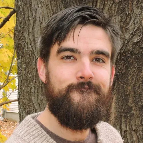 Man with a full beard smiles at the camera, wearing a light sweater, in front of a tree in autumn.