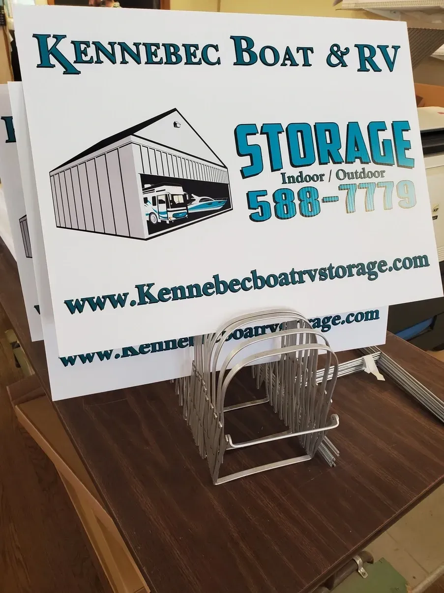 Signs for Kennebec Boat & RV Storage, white with teal lettering, in a metal holder on a brown table.
