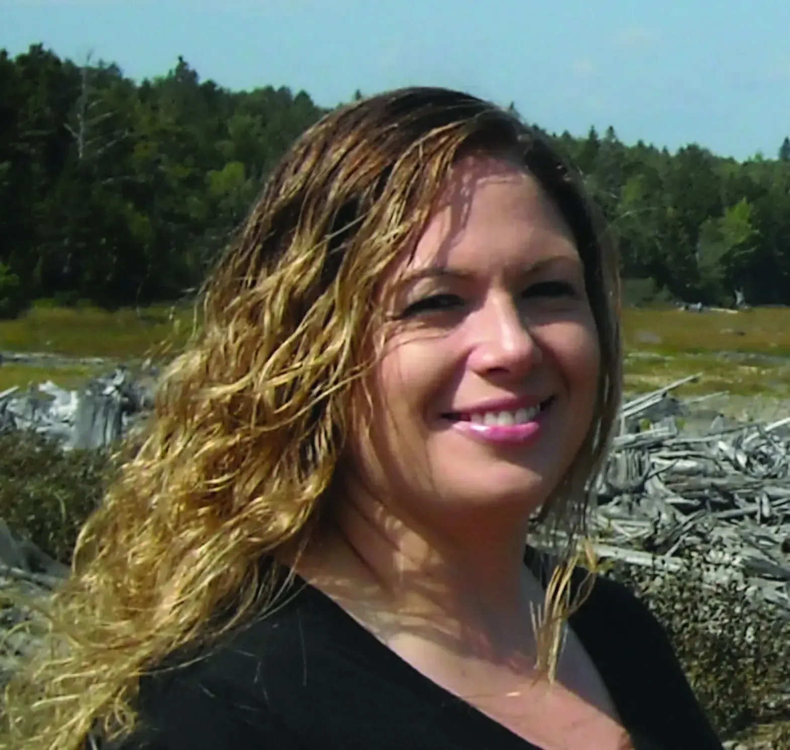 Woman with long, wavy blonde hair smiles outdoors. Background features trees and debris.