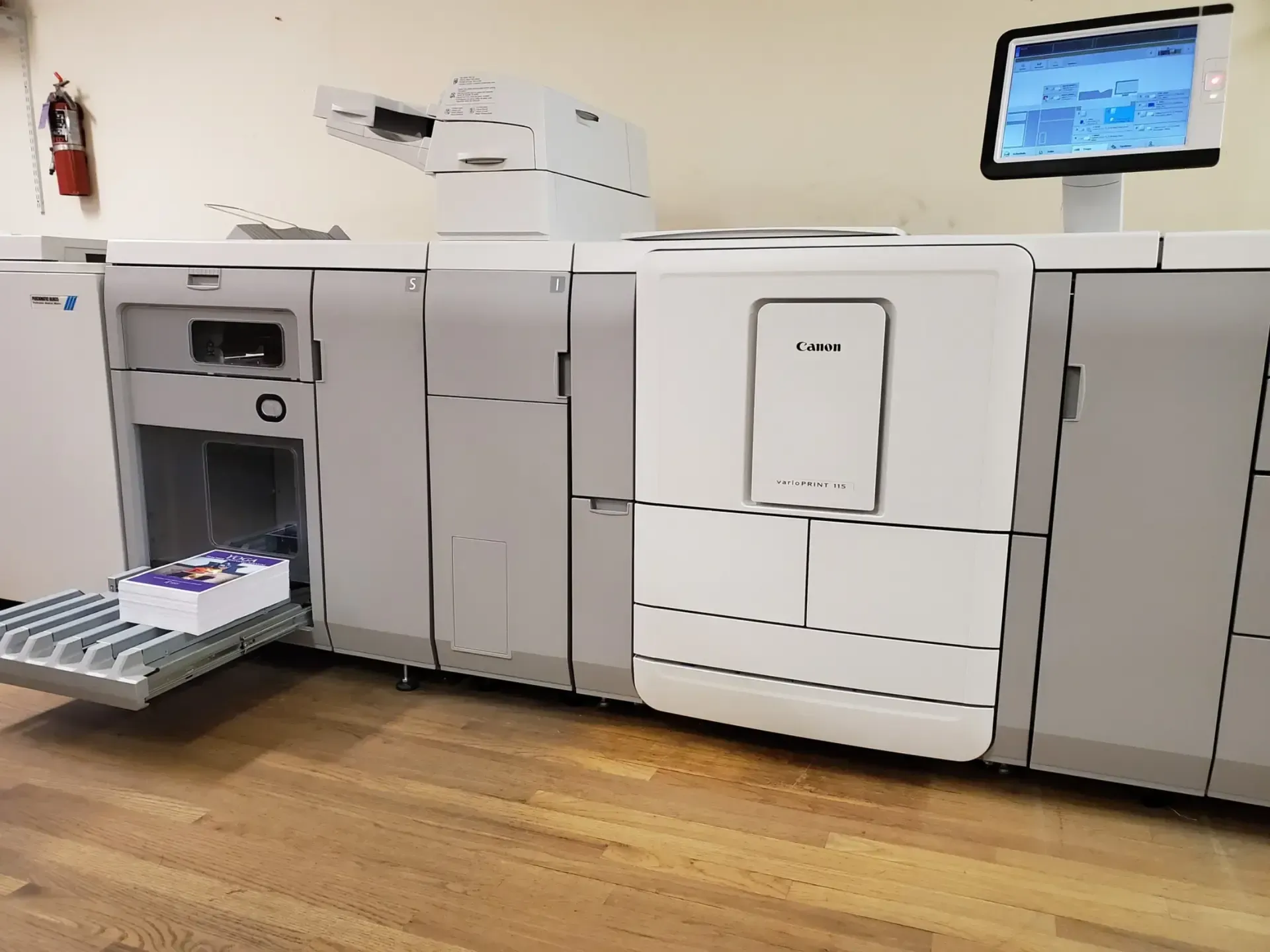Large digital printing machine with a stack of printed material on the output tray, a control panel, and a fire extinguisher on the wall.