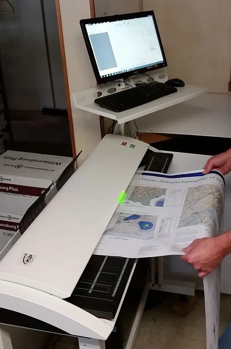 Person scans a large map with a wide scanner. A computer monitor sits above.