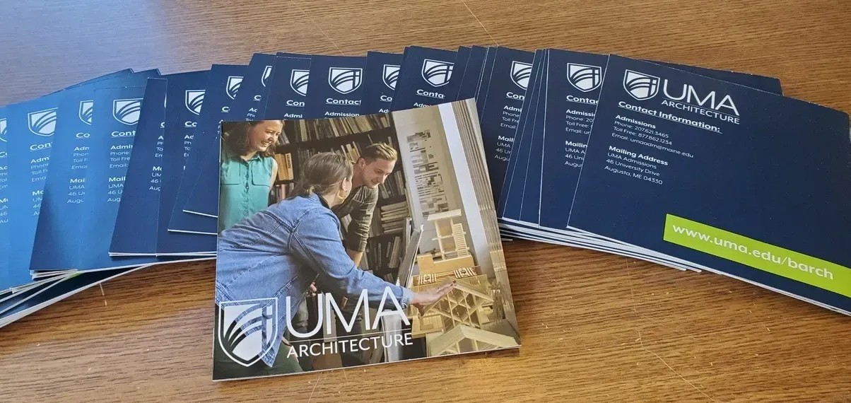 A stack of UMA Architecture brochures on a wooden table, with a cover photo of people looking at a model.
