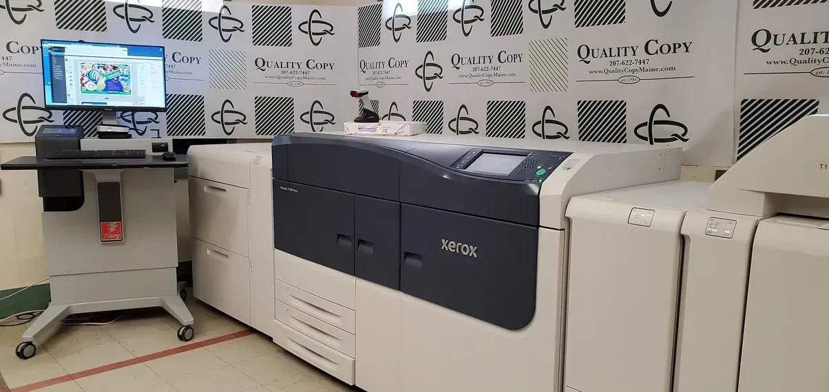 A large commercial printer in a print shop setting with a computer monitor and printer controls. The printer is beige and dark blue with a black and white patterned wall.