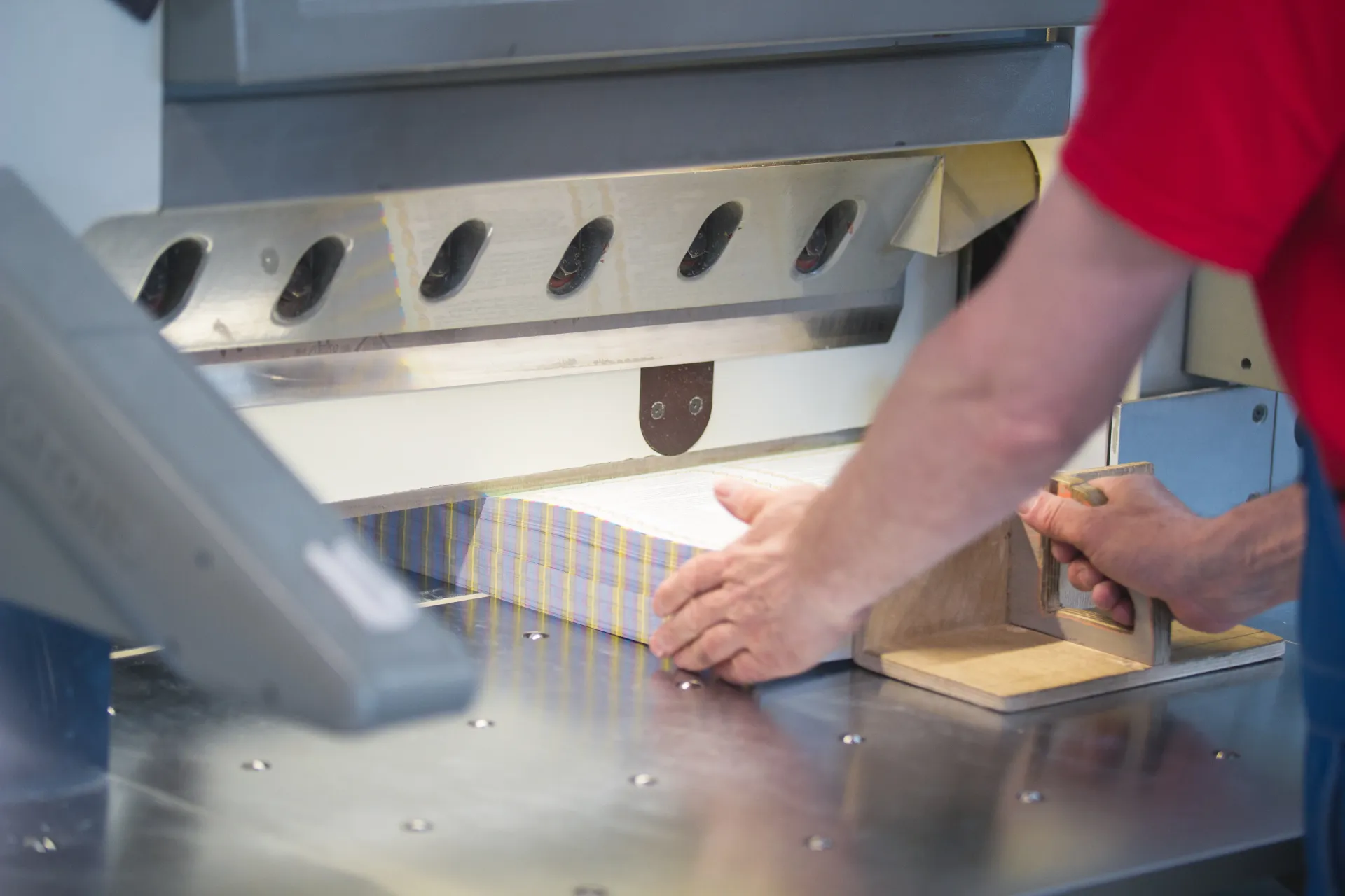 Person using a paper cutter to trim a stack of white paper on a metal table.