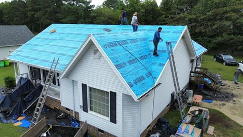 Three workers on a roof covered in bright blue underlayment, with ladders positioned against the siding of the house.