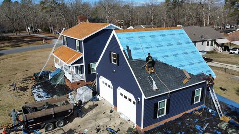 Roofers work on a navy blue house, replacing a roof with exposed blue underlayment and patches of new dark shingles.